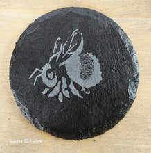 Load image into Gallery viewer, Round black slate coaster with engraved bee design on a wooden surface