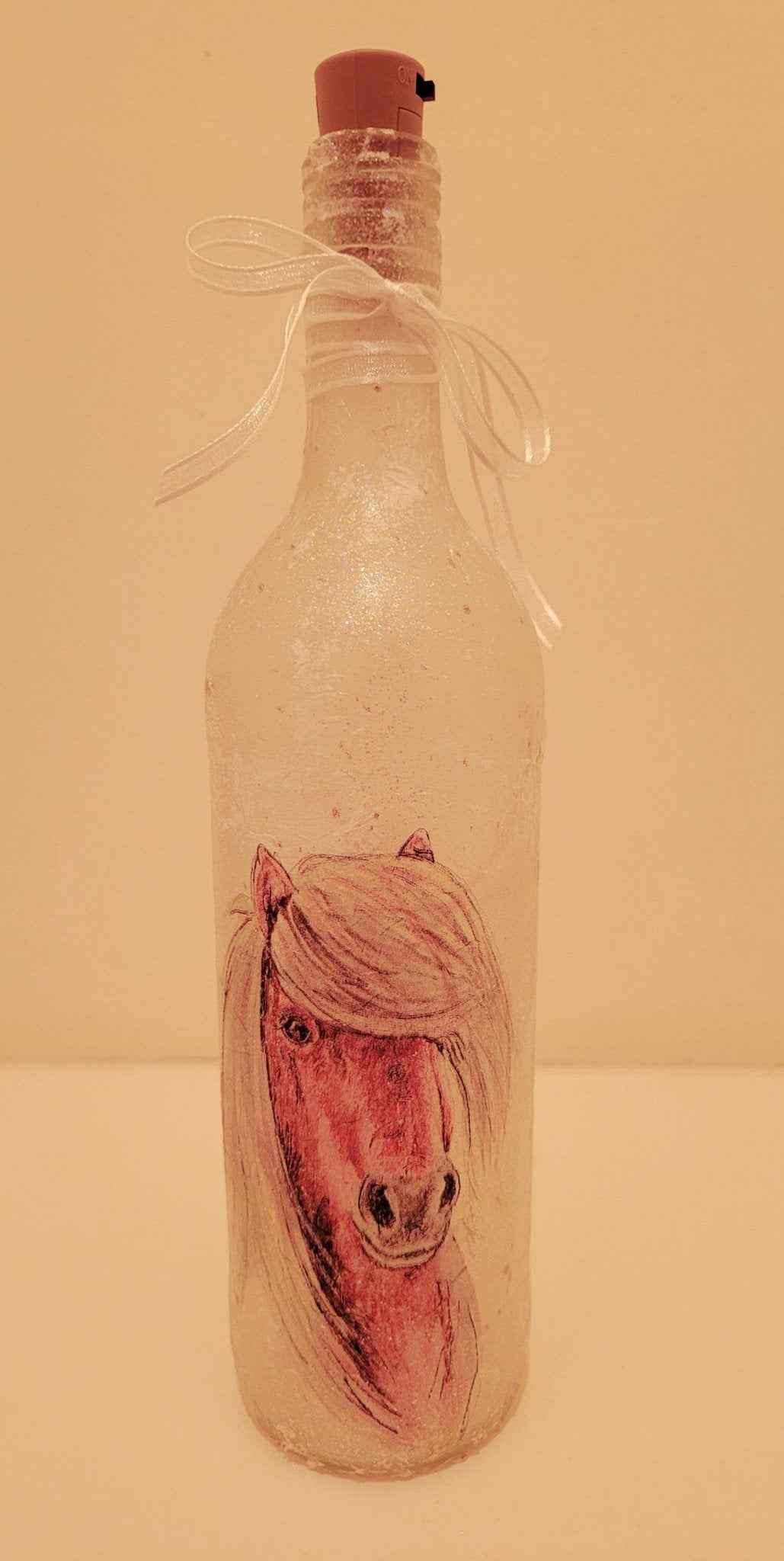 A frosted glass bottle with a cork stopper and a white ribbon bow, featuring a painted brown horse head on the front.