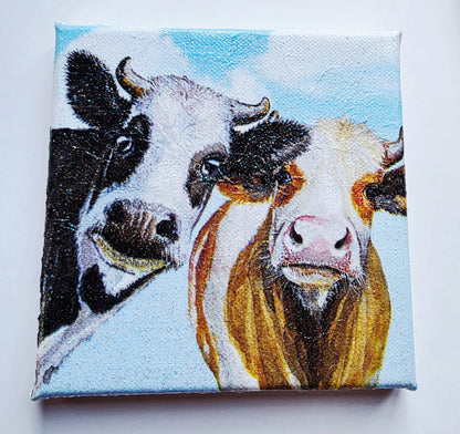 two cows decoupaged on stretched white canvas. One black and white, one brown and white looking nosy with blue sky background