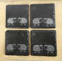 Load image into Gallery viewer, Four black slate coasters with white sheep illustrations on a wooden surface.

