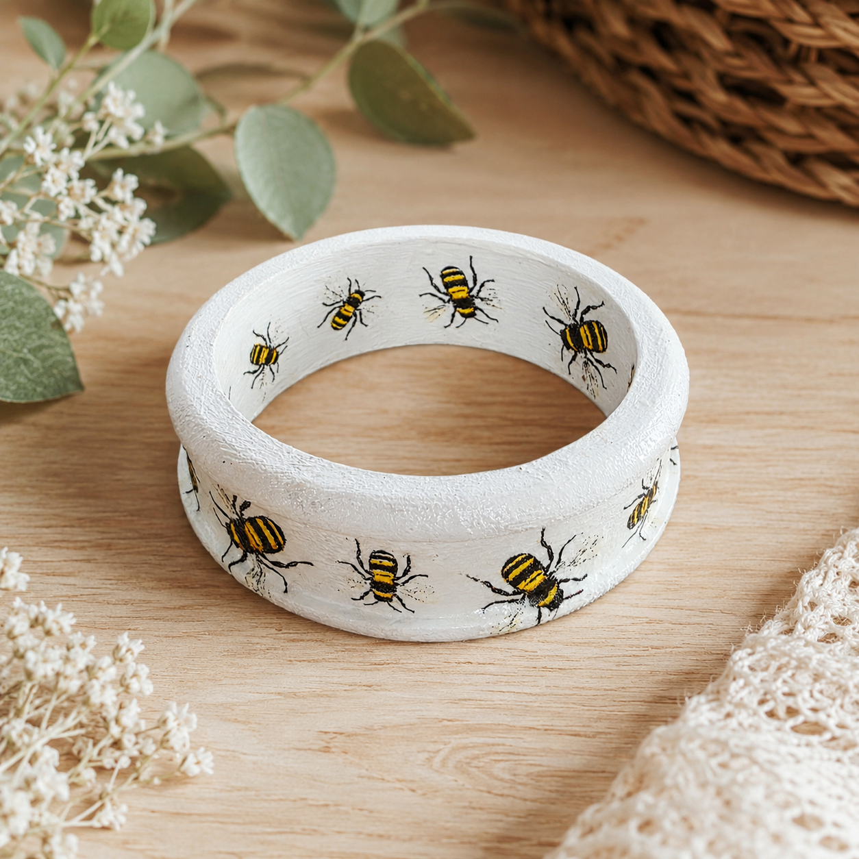 A white textured bangle bracelet decorated with painted yellow and black bumblebees, displayed on a light wooden surface with decorative green leaves and white flowers.