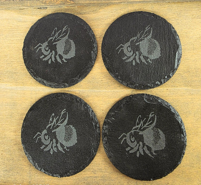 Four black slate coasters with engraved Bumble bee designs on a wooden surface