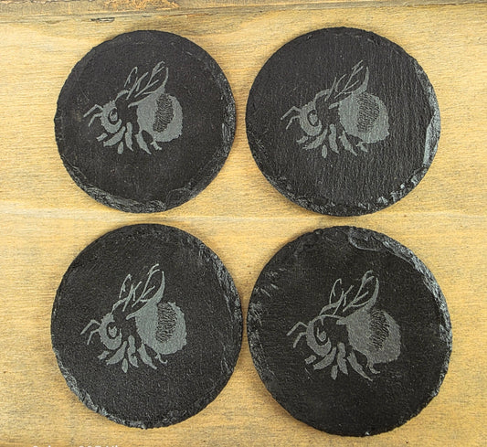 Four black slate coasters with engraved Bumble bee designs on a wooden surface