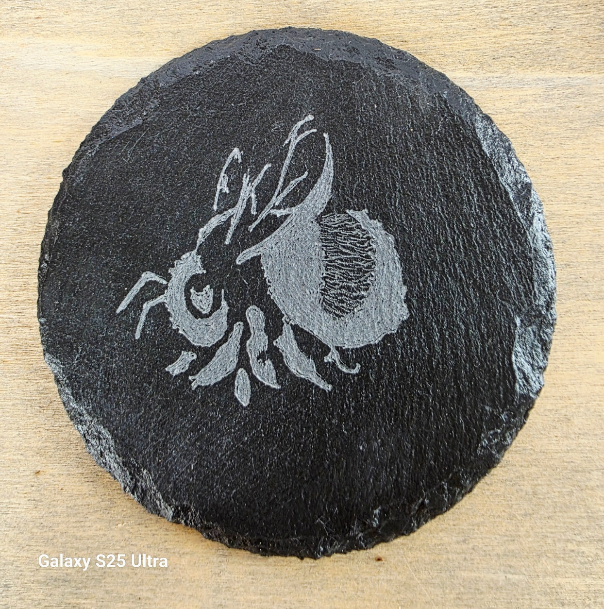Round black slate coaster with engraved bee design on a wooden surface