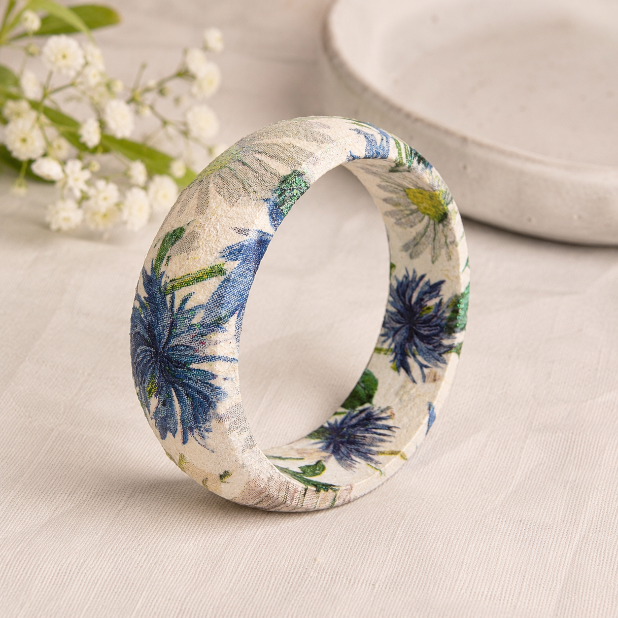 A bangle bracelet with a white base and a botanical pattern featuring green leaves, blue flowers, and a small yellow flower, and bee displayed on a cream background with asters nearby.