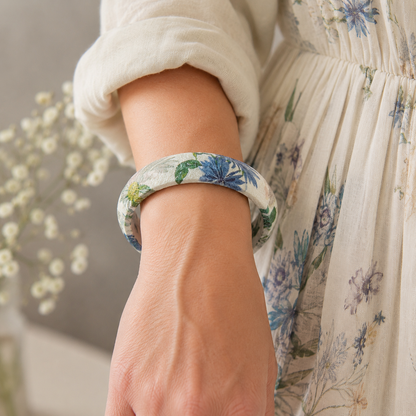 A close-up of a person's wrist wearing a wooden bangle bracelet with a blue and green floral print on a light background. The person is also wearing a cream-colored dress with a matching floral pattern.