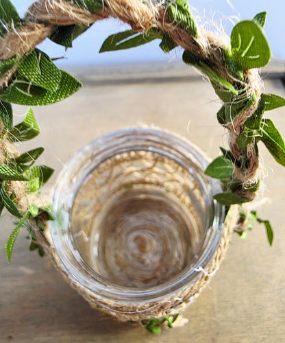 Glass jar with a decorative handle made of green leaves and twine against a light  background. Showing empty inside of jar