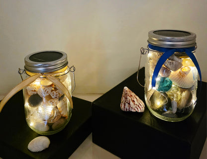 Two Jars filled with shells, pebbles and coloured glass stones. Lit up with lid and carry handle. Blue decorative ribbon