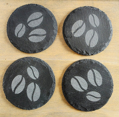 Four round black slate coasters with coffee bean design on a wooden surface