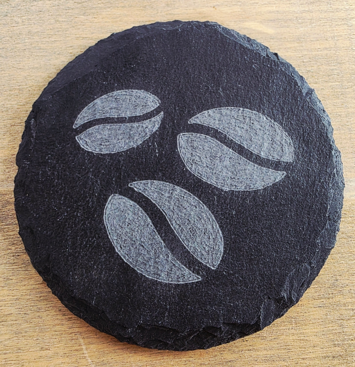 Round black slate coaster with engraved coffee bean design on a wooden surface