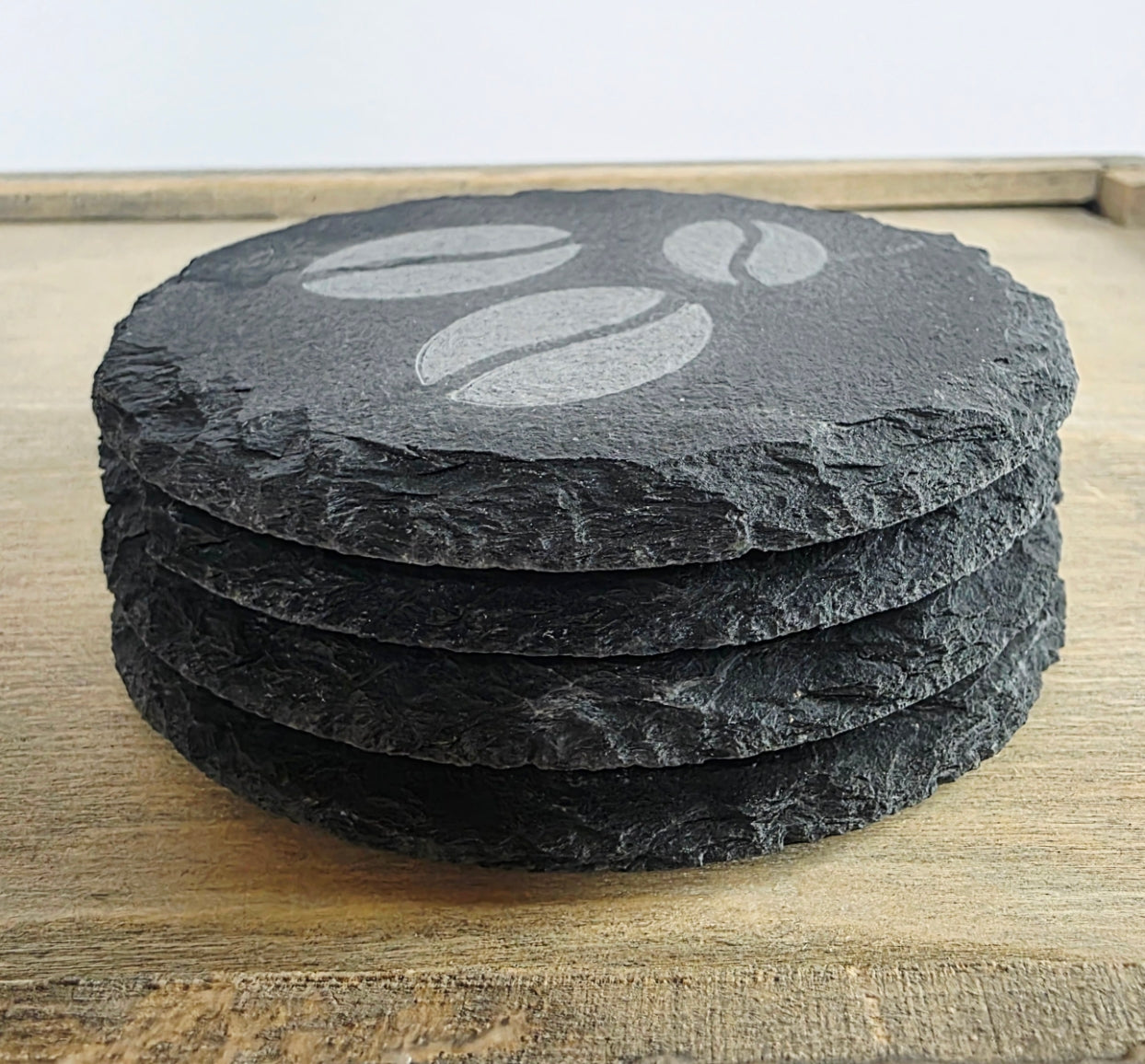 Stack of four round slate coasters with a coffee bean design on a wooden surface