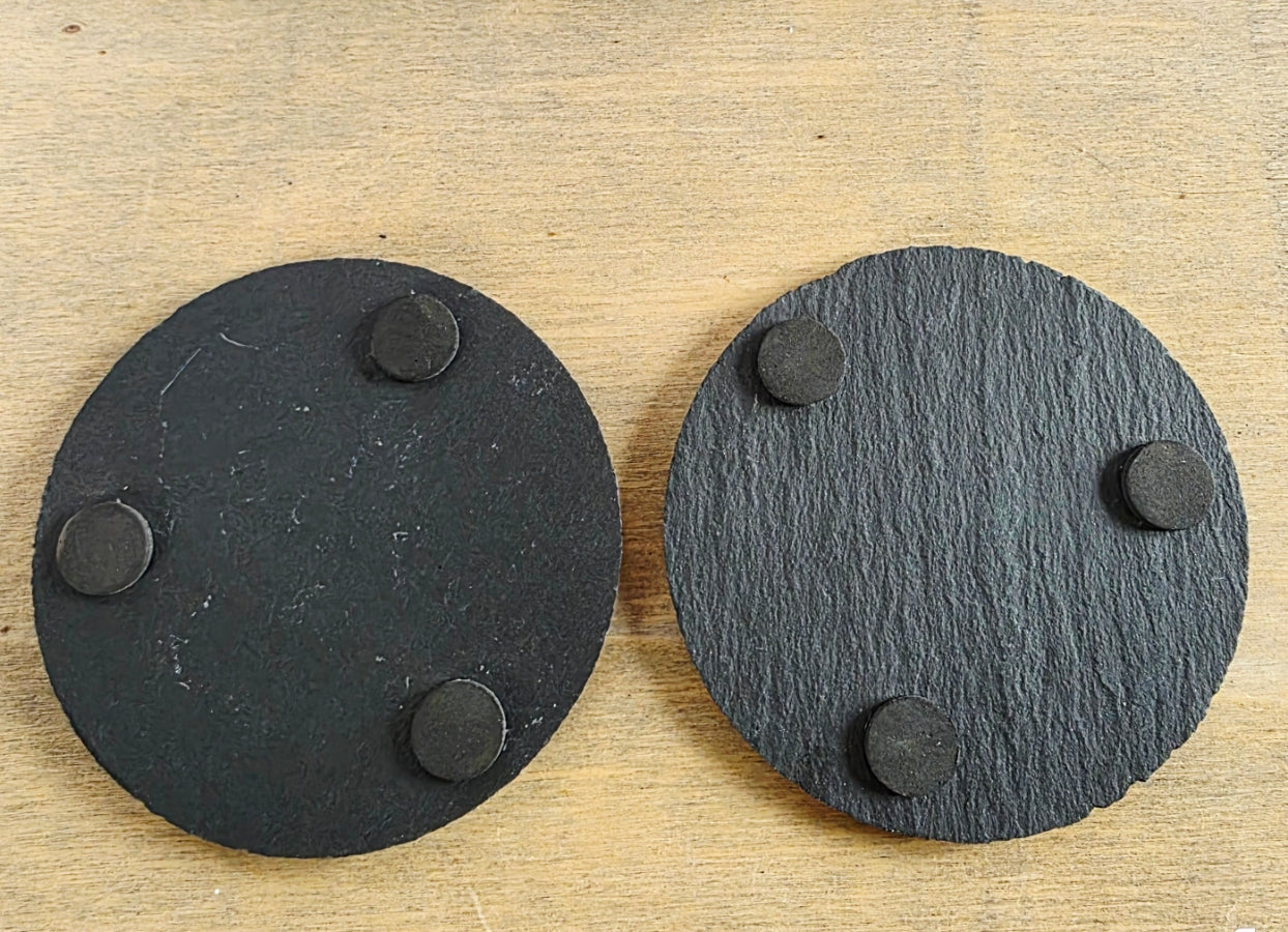 Two black circular coasters with three protective pads on the underside on each.