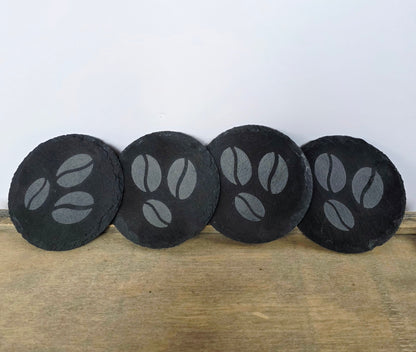 Four black, round slate coasters with coffee bean design on a wooden surface