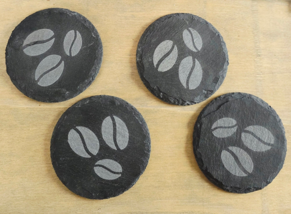 Four round black slate coasters with coffee bean design on a wooden surface