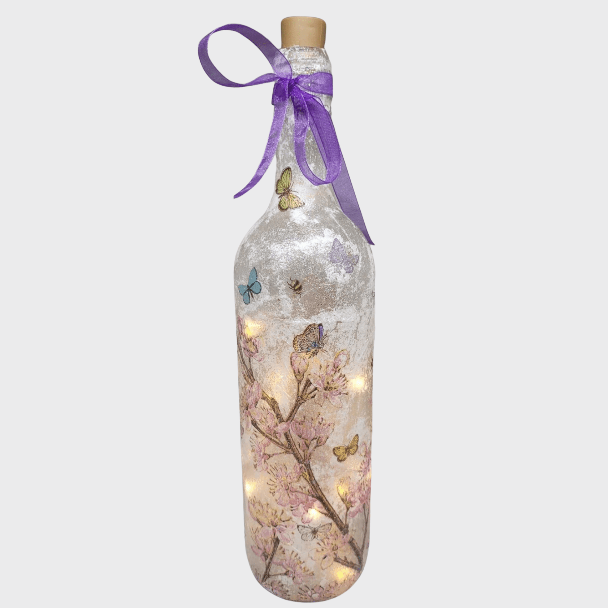 Decoupage Flower Butterfly Bottle Lamp - standard wine bottle with pink blossom detail and yellow/blue/reen butterflies. Purple ribbon tied around the neck of the bottle. Lit up with cork LED lights