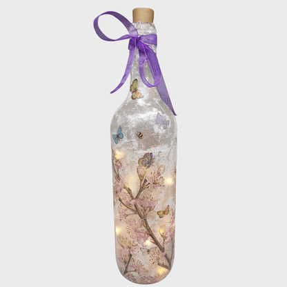 Decoupage Flower Butterfly Bottle Lamp - standard wine bottle with pink blossom detail and yellow/blue/reen butterflies. Purple ribbon tied around the neck of the bottle. Lit up with cork LED lights