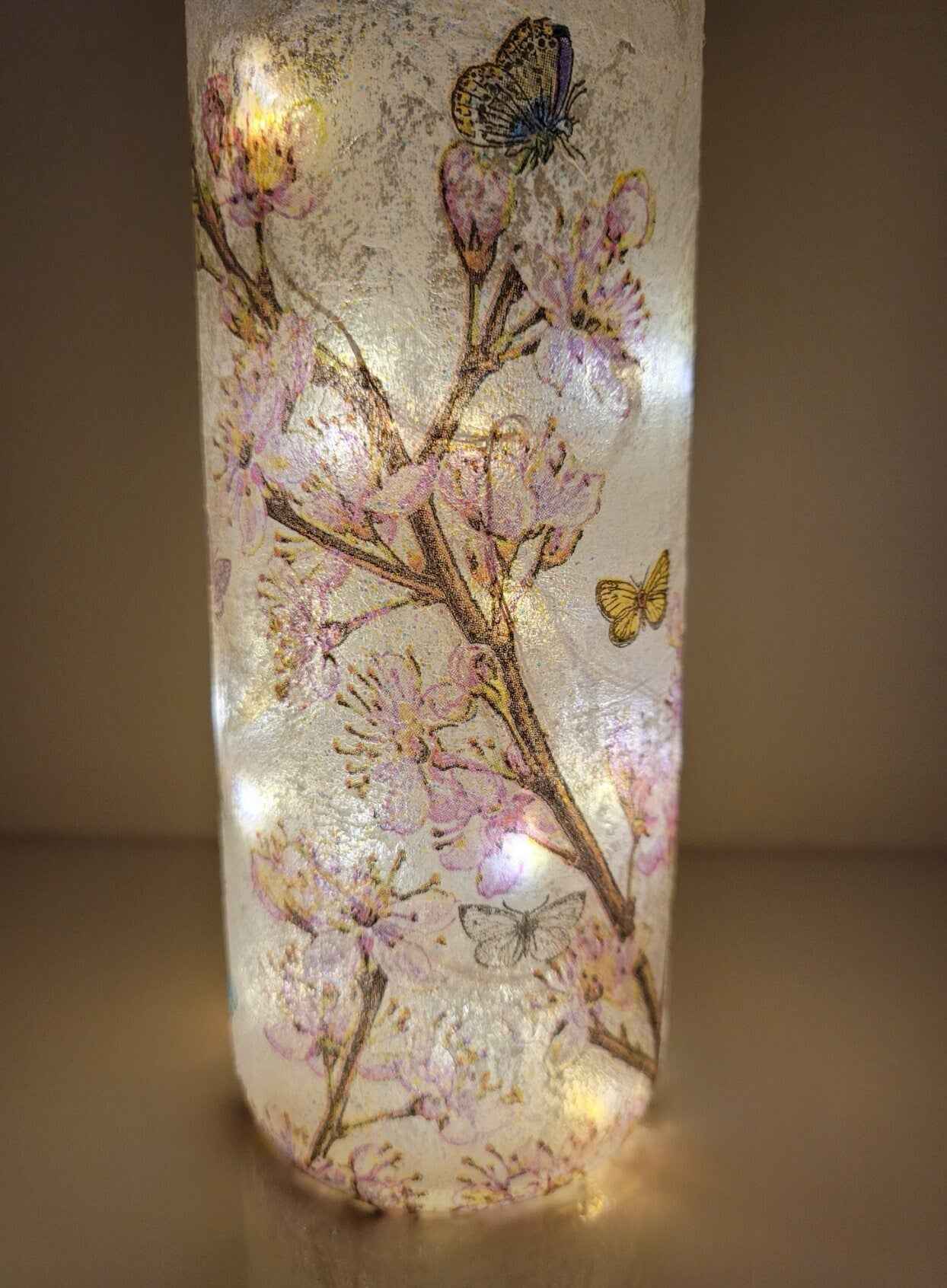 A standard wine bottle with a warm white glow from internal string lights, decorated with pressed pink cherry blossoms and butterflies on a textured glass surface.