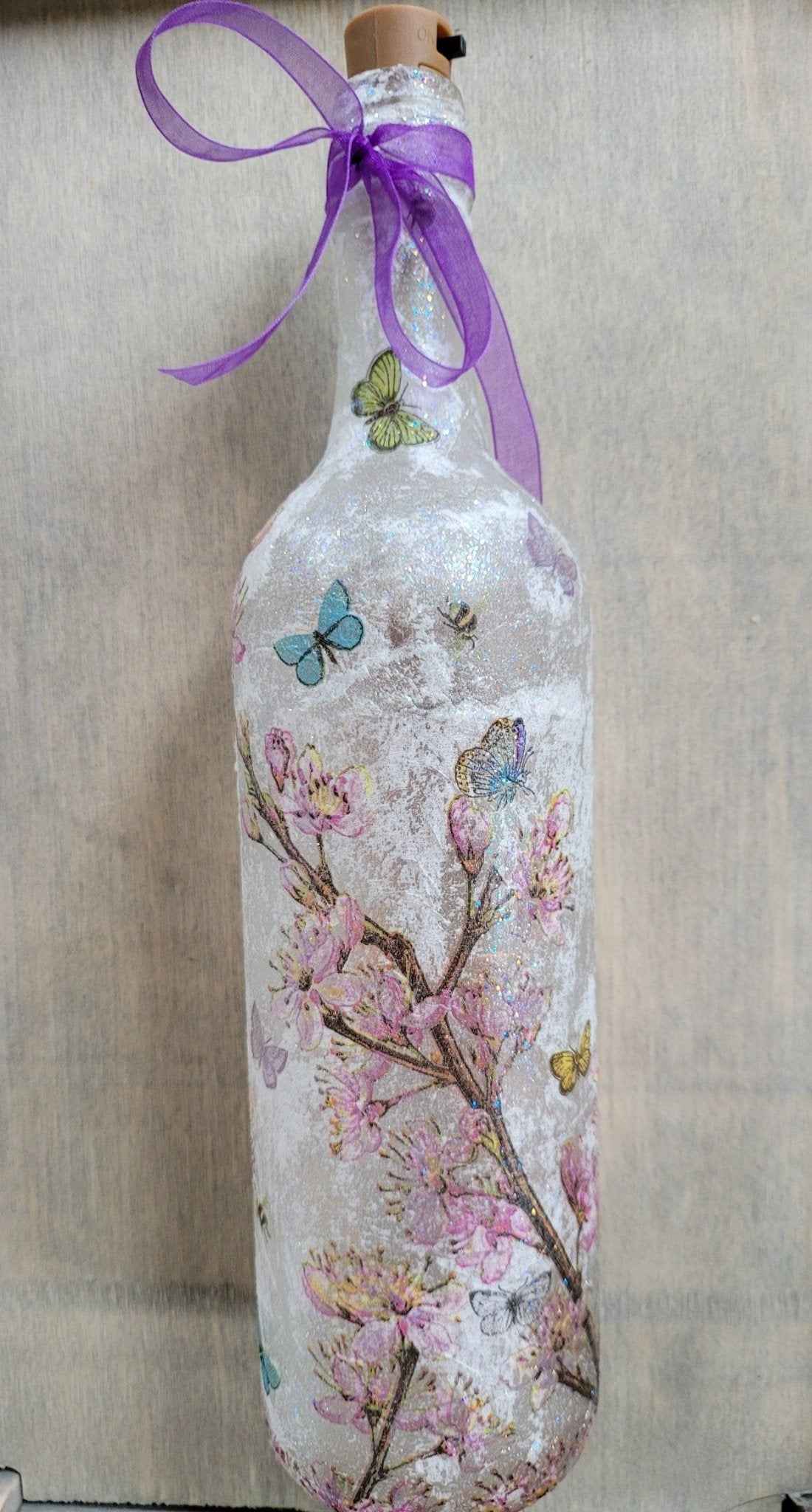 A frosted glass bottle with a cork stopper, decorated with pink cherry blossom branches, blue butterflies, and a purple ribbon bow at the neck, set against a neutral beige fabric background.
