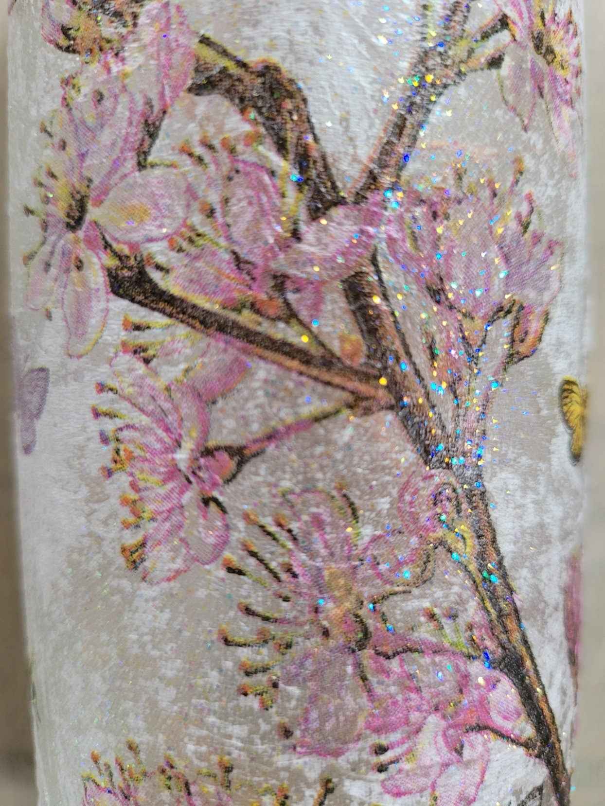 A close-up of a standard wine bottle featuring a textured off-white surface decorated with pink cherry blossom flowers and brown branches, accented with iridescent glitter.
