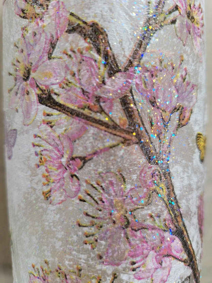 A close-up of a standard wine bottle featuring a textured off-white surface decorated with pink cherry blossom flowers and brown branches, accented with iridescent glitter.