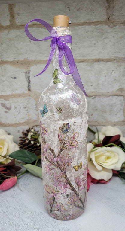 A clear glass bottle with a cork stopper, decorated with pink cherry blossom branches, butterflies, and a purple ribbon bow, set against a light brick wall with white roses in the background.