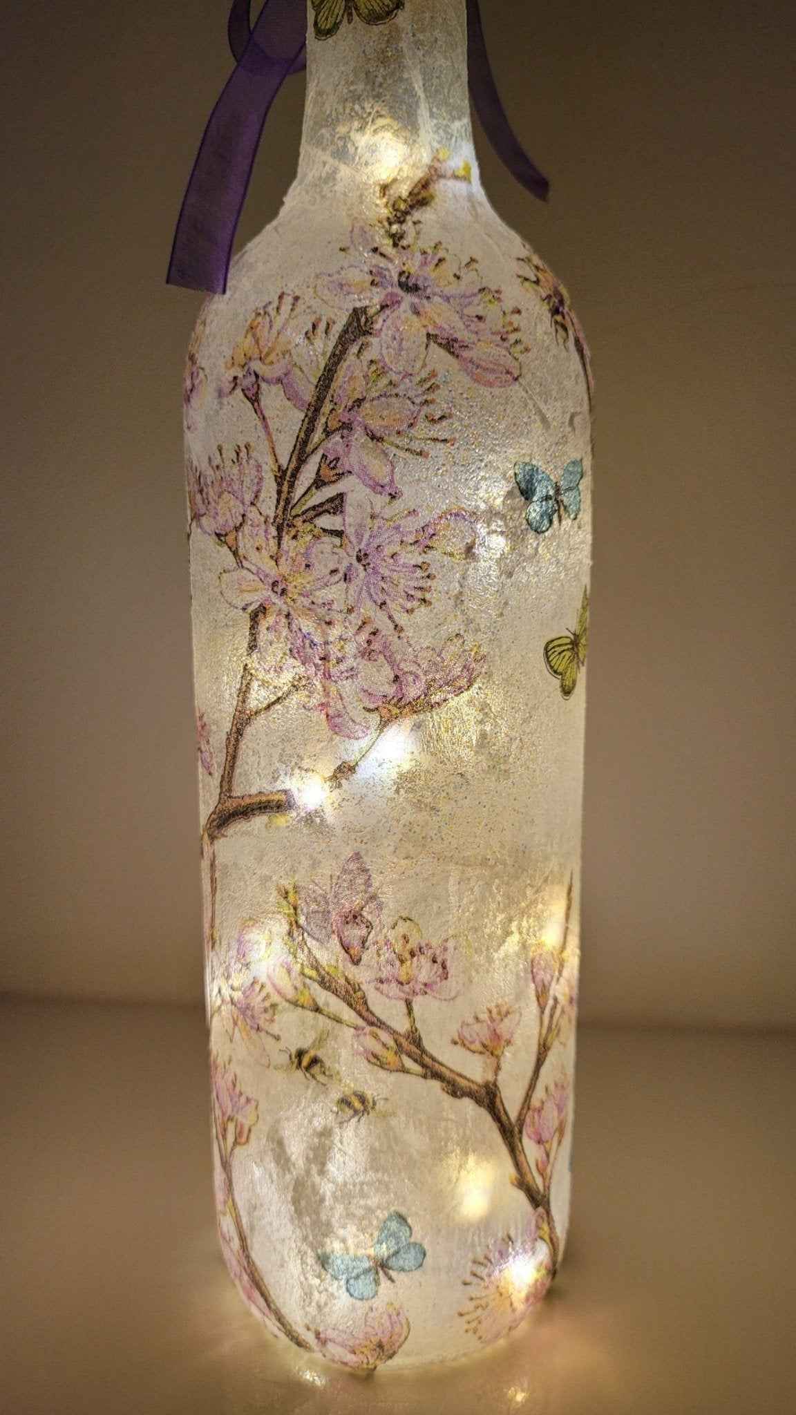 Decoupage Flower Butterfly Bottle Lamp - standard wine bottle with pink blossom detail and yellow/blue/reen butterflies. Purple ribbon tied around the neck of the bottle. Lit up with cork LED lights