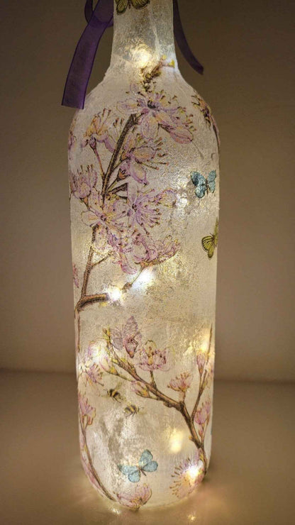 Decoupage Flower Butterfly Bottle Lamp - standard wine bottle with pink blossom detail and yellow/blue/reen butterflies. Purple ribbon tied around the neck of the bottle. Lit up with cork LED lights