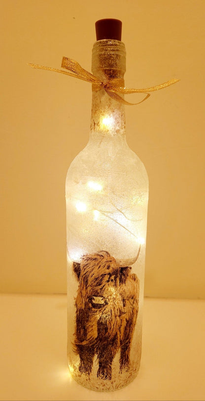 Lit up standard shape wine bottle with highland cow detailing on the front near the bottom. Gold ribbon tied around the neck of the bottle. Cork LED lights