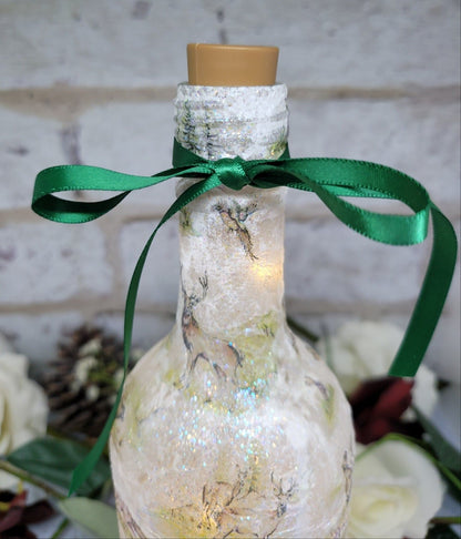 A decorative wine bottle with a woodland scene design. The bottle is illuminated with fairy lights and features a cork stopper and a dark green ribbon. The bottle's surface is textured and covered with images of deer, trees, and other forest animals.