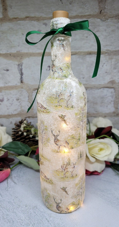 A decorative wine bottle with a woodland scene design. The bottle is illuminated with fairy lights and features a cork stopper and a dark green ribbon. The bottle's surface is textured and covered with images of deer, trees, and other forest animals.