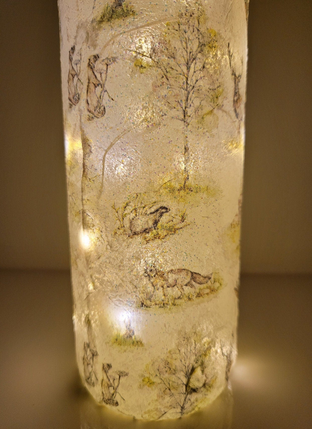 A decorative wine bottle with a woodland scene design. The bottle is illuminated with fairy lights and features a cork stopper and a dark green ribbon. The bottle's surface is textured and covered with images of deer, trees, and other forest animals.