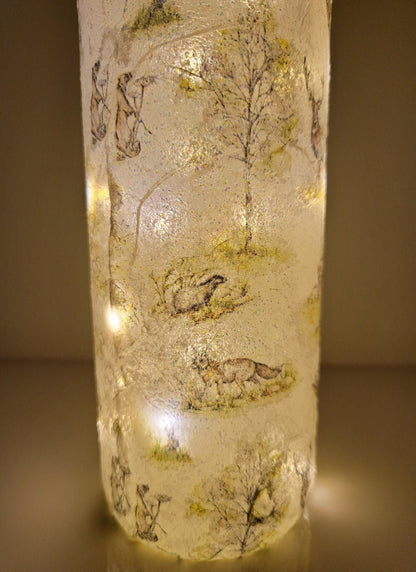 A decorative wine bottle with a woodland scene design. The bottle is illuminated with fairy lights and features a cork stopper and a dark green ribbon. The bottle's surface is textured and covered with images of deer, trees, and other forest animals.