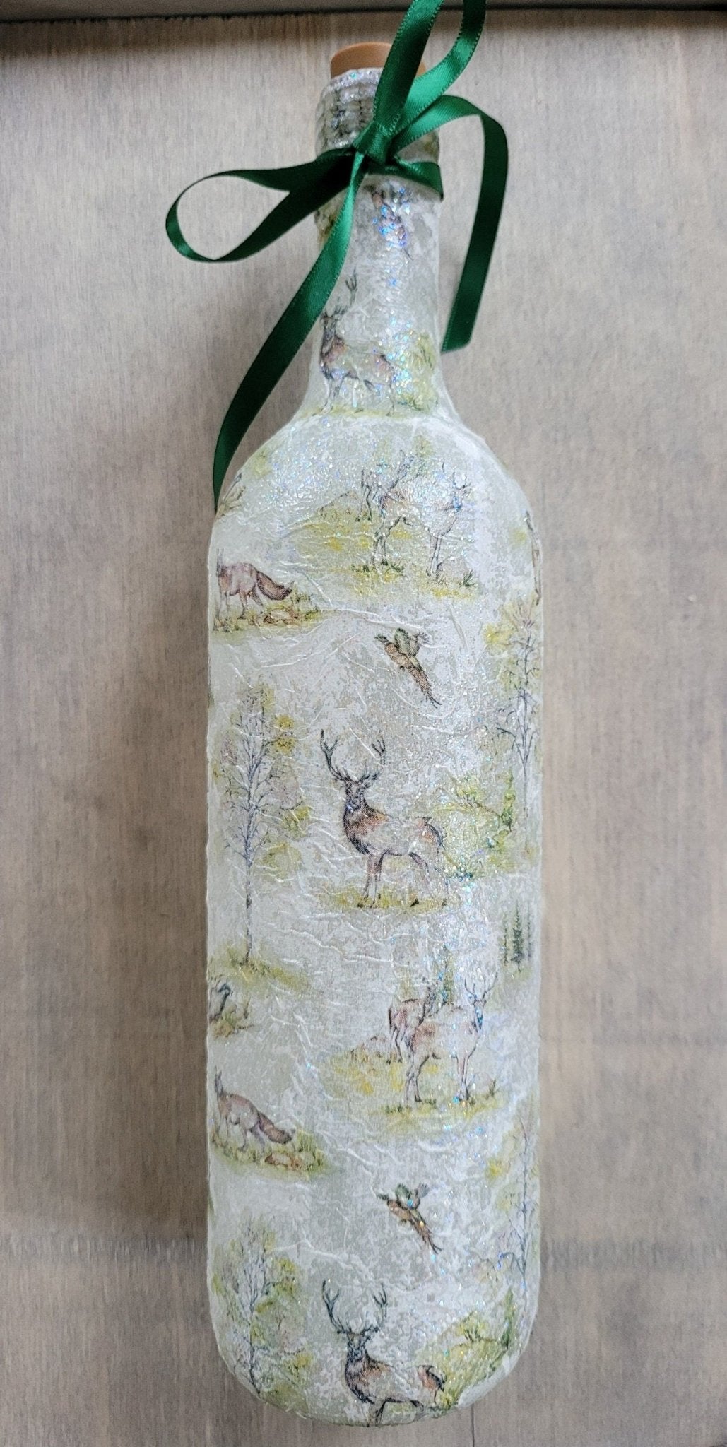 A decorative wine bottle with a woodland scene design. The bottle is illuminated with fairy lights and features a cork stopper and a dark green ribbon. The bottle's surface is textured and covered with images of deer, trees, and other forest animals.