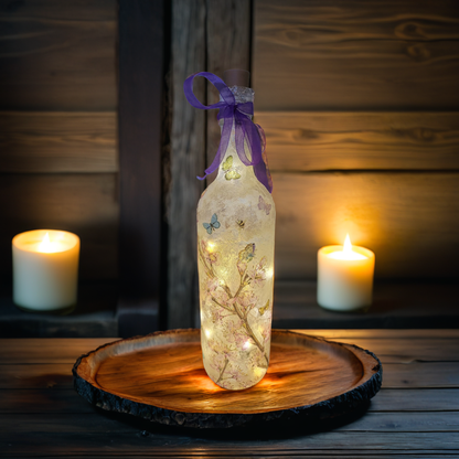 A decoupage flower and butterfly decorated glass bottle lamp with a purple satin bow, lit and placed on a wooden tray with candles in the background.
