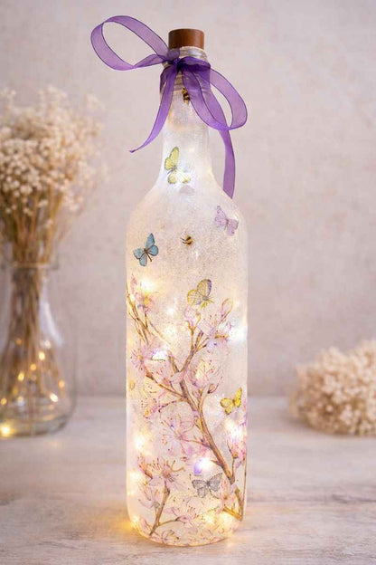 A decorative clear glass bottle with a cork stopper and a purple ribbon bow, filled with warm fairy lights and decorated with pressed cherry blossoms and butterflies. The bottle is photographed on a tabletop with soft-focus dried flowers in the background.