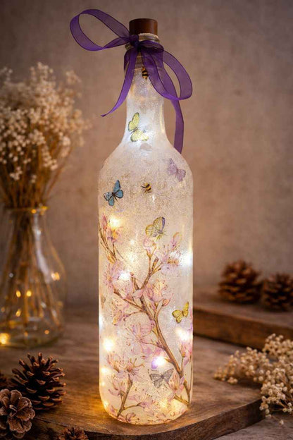 A clear glass bottle with a cork stopper, decorated with pressed cherry blossom branches, butterflies, and warm white fairy lights inside, tied with a purple ribbon. The bottle is styled on a wooden surface with pinecones and dried flowers in the background.