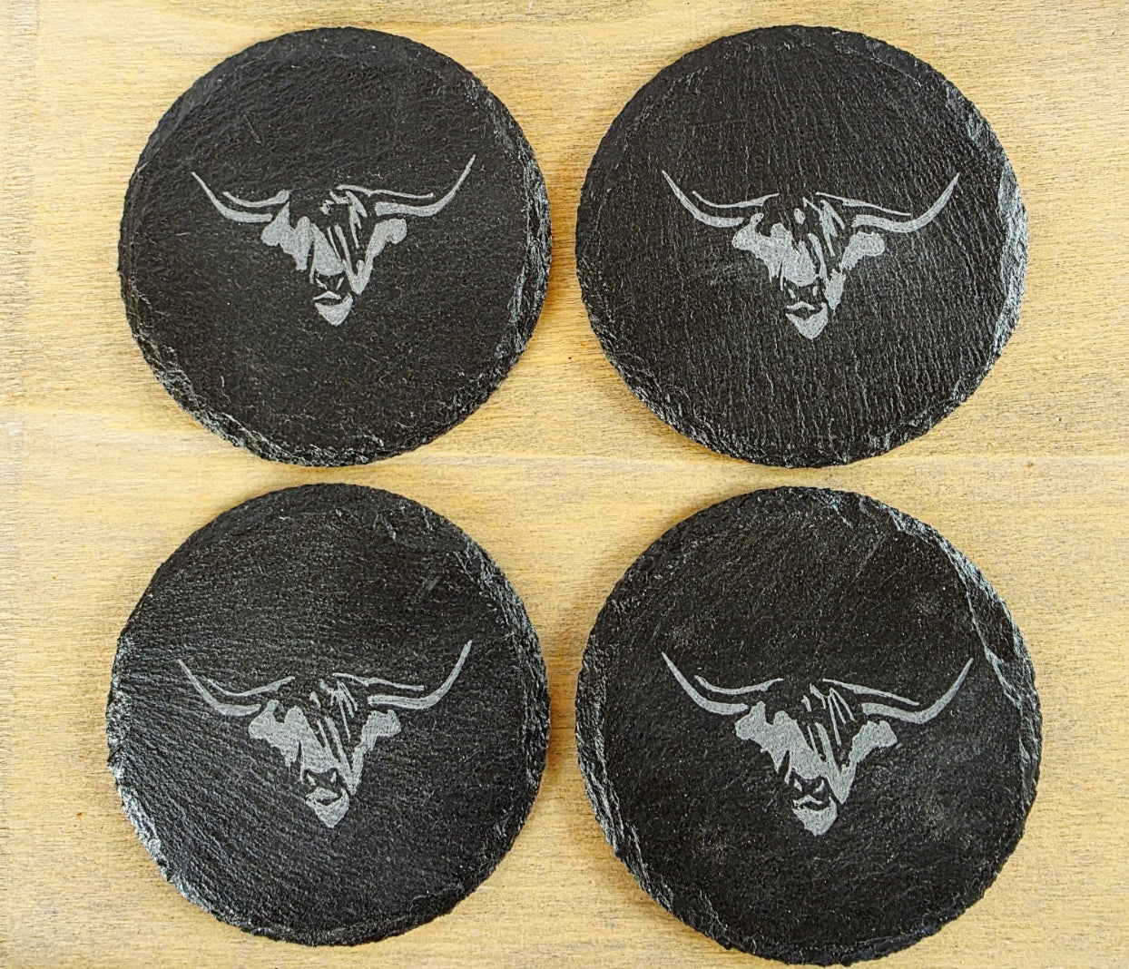 Four black slate coasters with cow's head design on a wooden surface