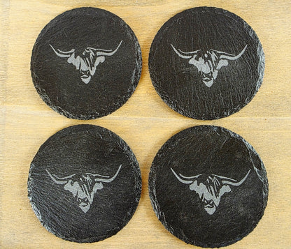 Four black slate coasters with cow's head design on a wooden surface