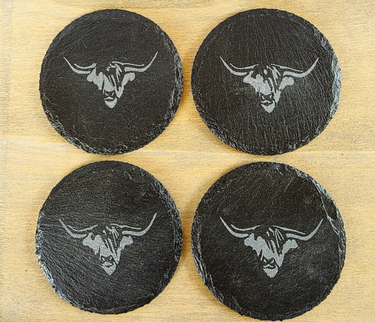 Four black slate coasters with cow's head design on a wooden surface