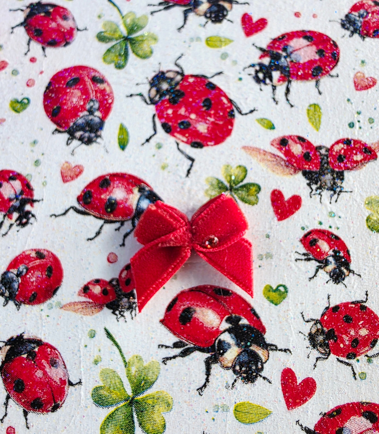 Pattern of red ladybugs, green leaves, and red hearts on a white background with a red bow.
