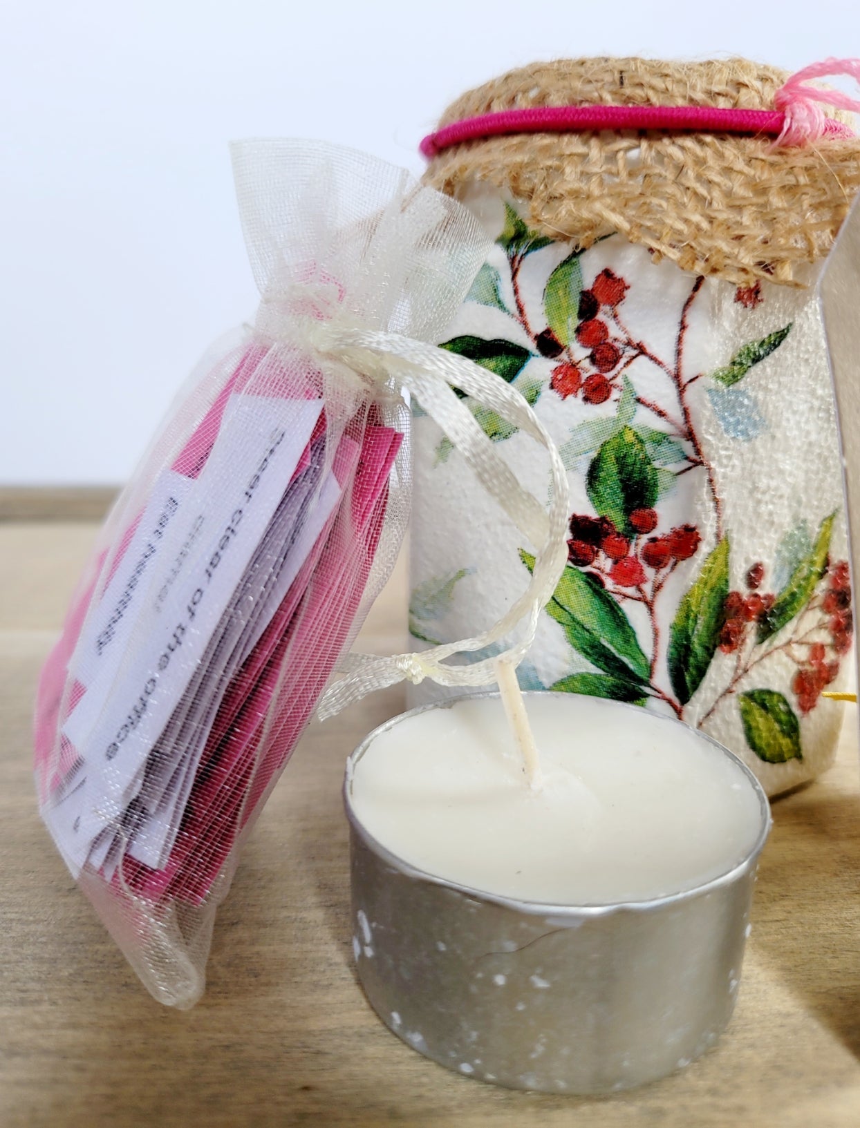 Hessian lid topped jar showing contents outside; pale organza bag with white/pink quotes, and a tealight candle. Jar s white painted with a red and green berry leaf design