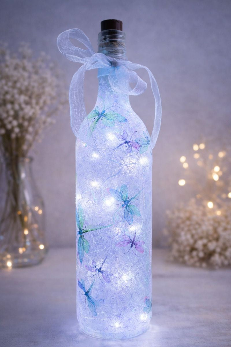 Decorative bottle with LED lights and dragonfly designs, surrounded by blurred flowers and lights.