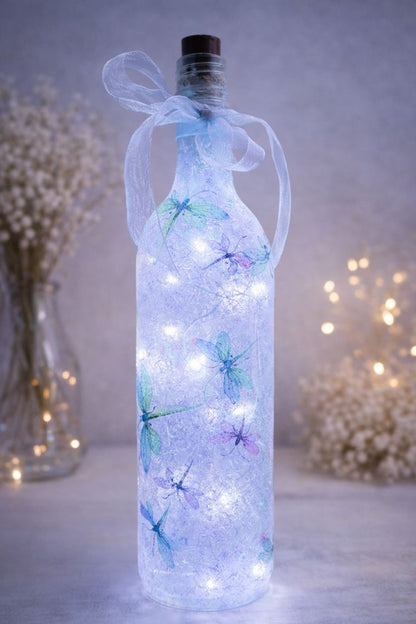Decorative bottle with LED lights and dragonfly designs, surrounded by blurred flowers and lights.