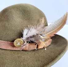 Load image into Gallery viewer, Pheasant feather hat pin, long pheasant tail feather with shorter feathers on the end of a brass gold coloured shotgun cartridge. Shown pinned onto the side of a green trilby style hat
