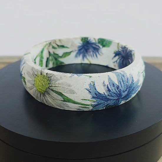 A bangle bracelet with a white base and a botanical pattern featuring green leaves, blue flowers, and a small yellow flower, and bee displayed on a black rotating stand