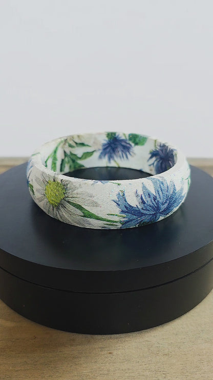 A bangle bracelet with a white base and a botanical pattern featuring green leaves, blue flowers, and a small yellow flower, and bee displayed on a black rotating stand