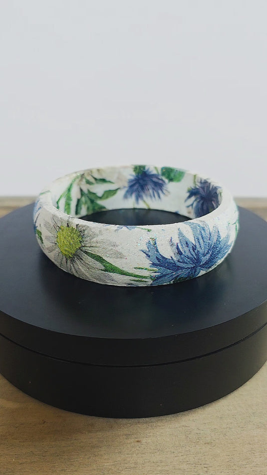 A bangle bracelet with a white base and a botanical pattern featuring green leaves, blue flowers, and a small yellow flower, and bee displayed on a black rotating stand