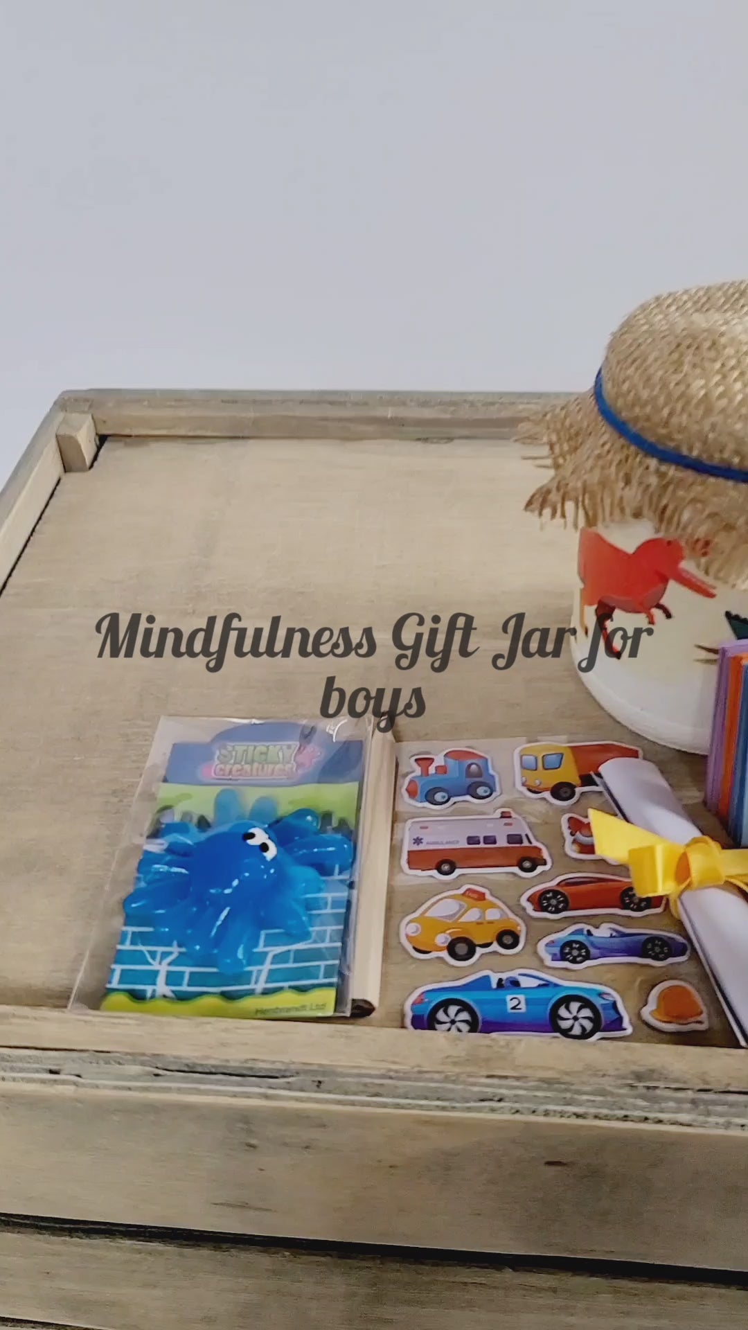 Video of a White painted jar with Dinosuar detail. Hessian lid secured with a blue band, sticker sheet, blue sticky toy, lit up flameless votive candle, white rolled up note secured with yellow ribbon, key ring attached to jar with a green Dinosaur on it. Coloured card scrapbook and short brown pencil, organza bag containing pink and blue quotes