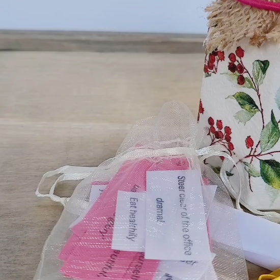 video showing a small white jar decorated with red berries and leaves, an organza bag with white and paper quotes inside. Jar has hessian lid in light brown, small white tealight candle and a white rolled up pice of paper tied with yellow ribbon