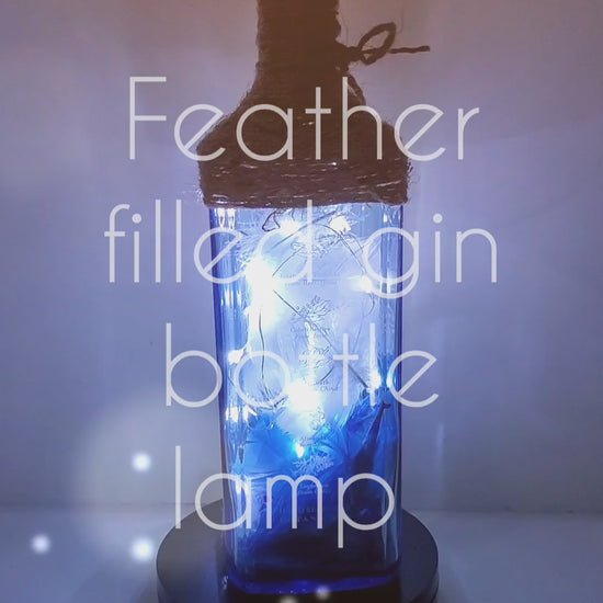 Rotating example of a Lit up blue glass gin bottle with jute twine detail around the top of the lamp, cork LED lights. Bottle is filled with blue and white feathers and blue coloured stones in the bottom.